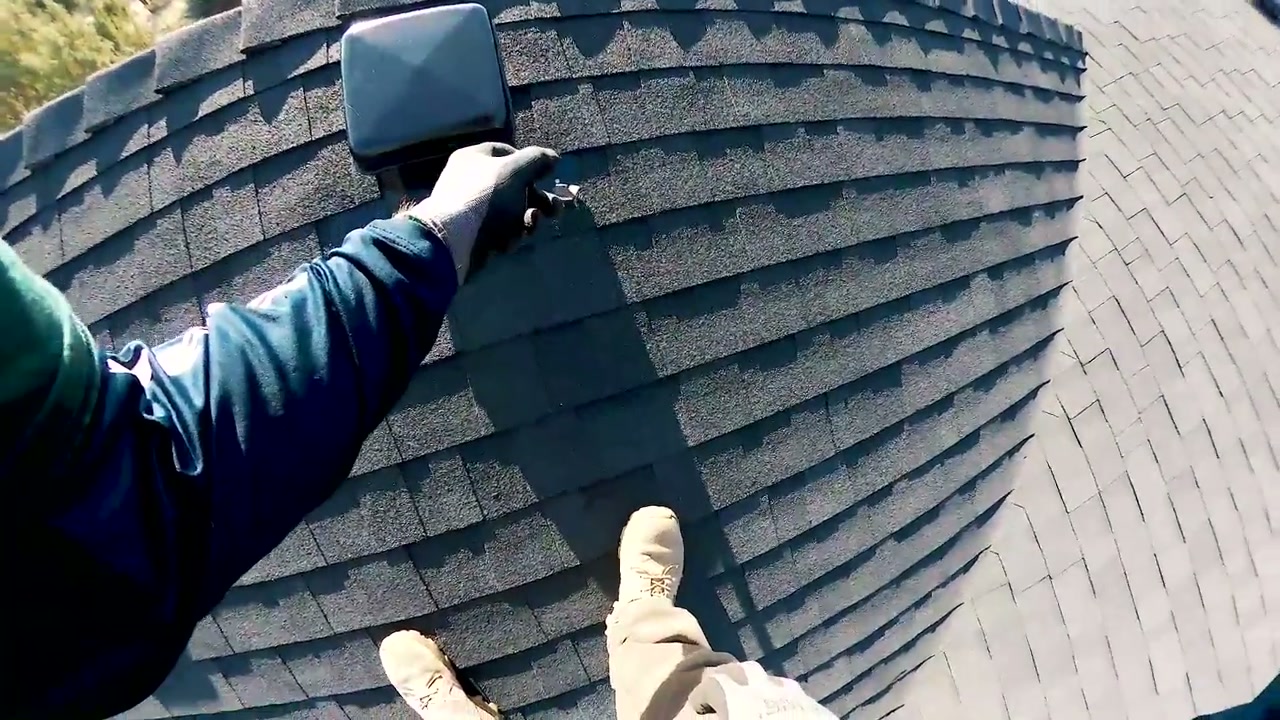 An inspector checks a fastening detail beside a roof vent on a shingle roof.